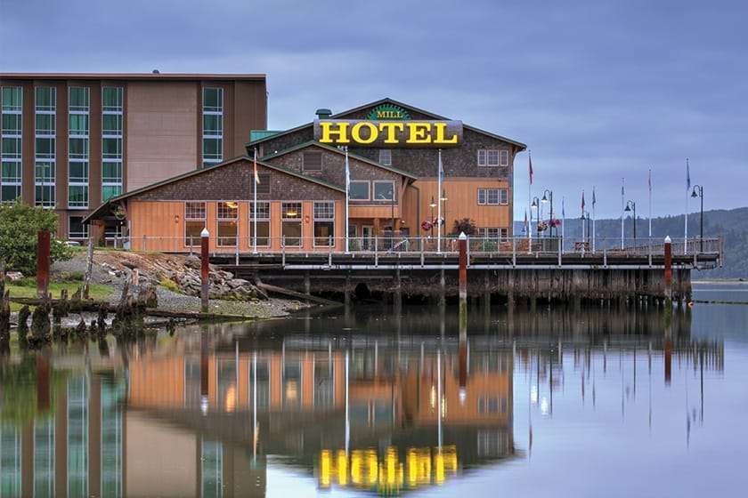 Brown and red hotel building on the bay. Yellow "Mill Hotel" letters on building. Building can be seen reflected in the surrounding water.